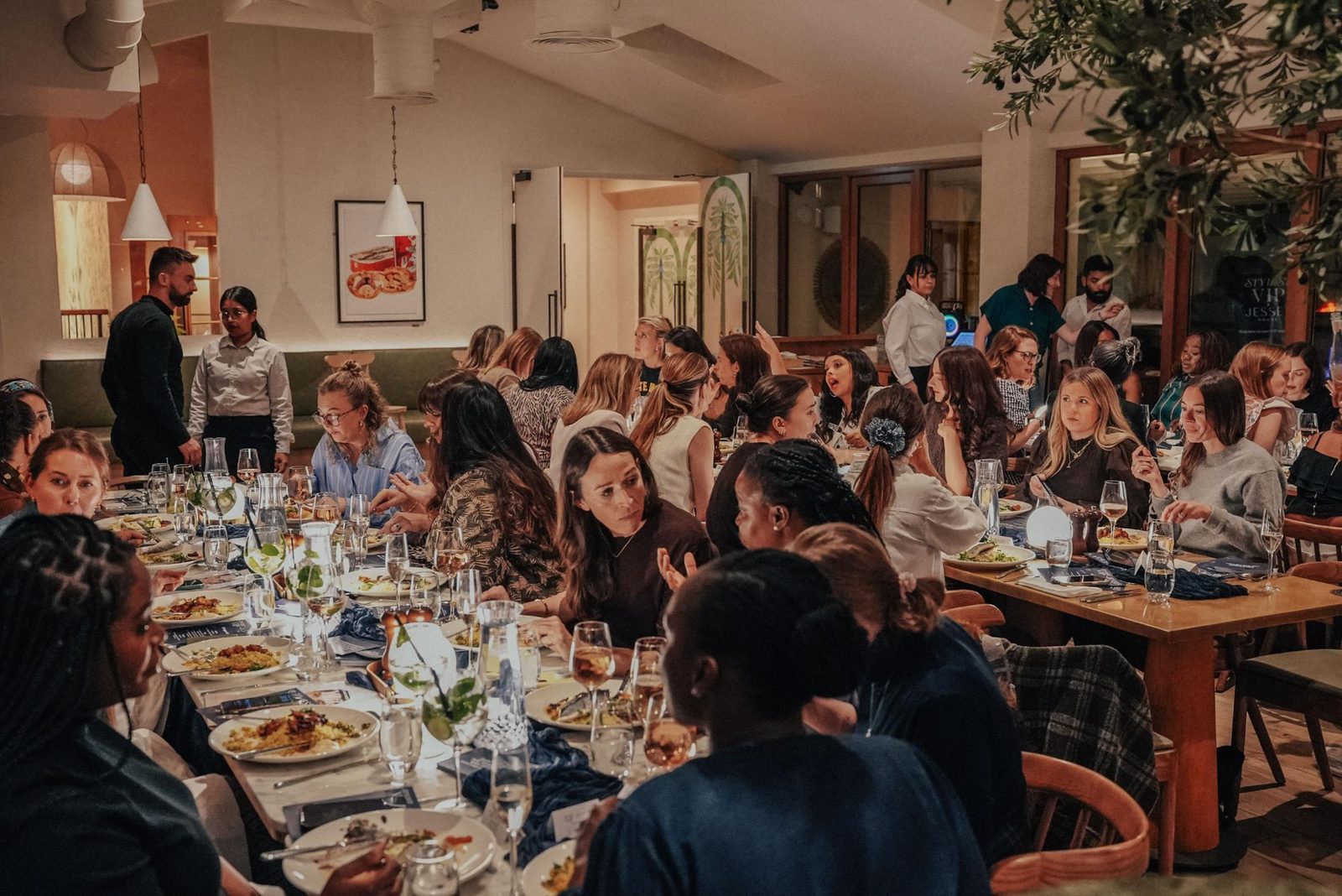 A lively restaurant scene with two long tables of people dining, talking, and enjoying food and drinks. Warm lighting and modern decor create a cozy, welcoming atmosphere.