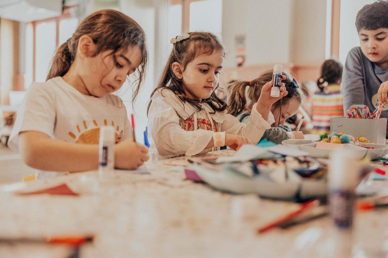 Several young children sit at a table engaged in arts and crafts, using glue sticks, markers, and colorful materials, focused on their creative projects in a bright, indoor classroom setting.