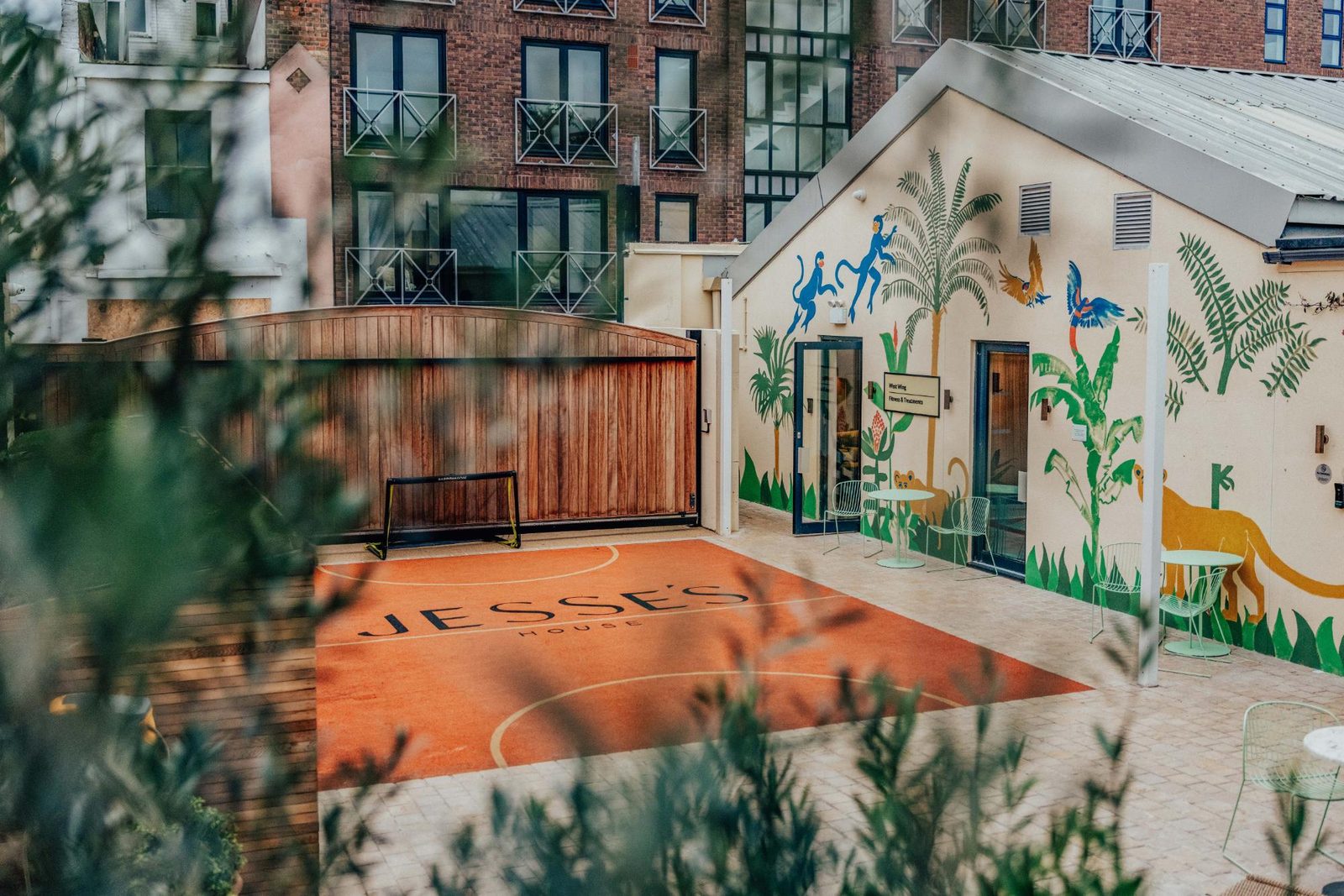 A small outdoor basketball court labeled Jesses House is surrounded by greenery. The adjacent building features colorful, playful animal murals. Apartment buildings are visible in the background.