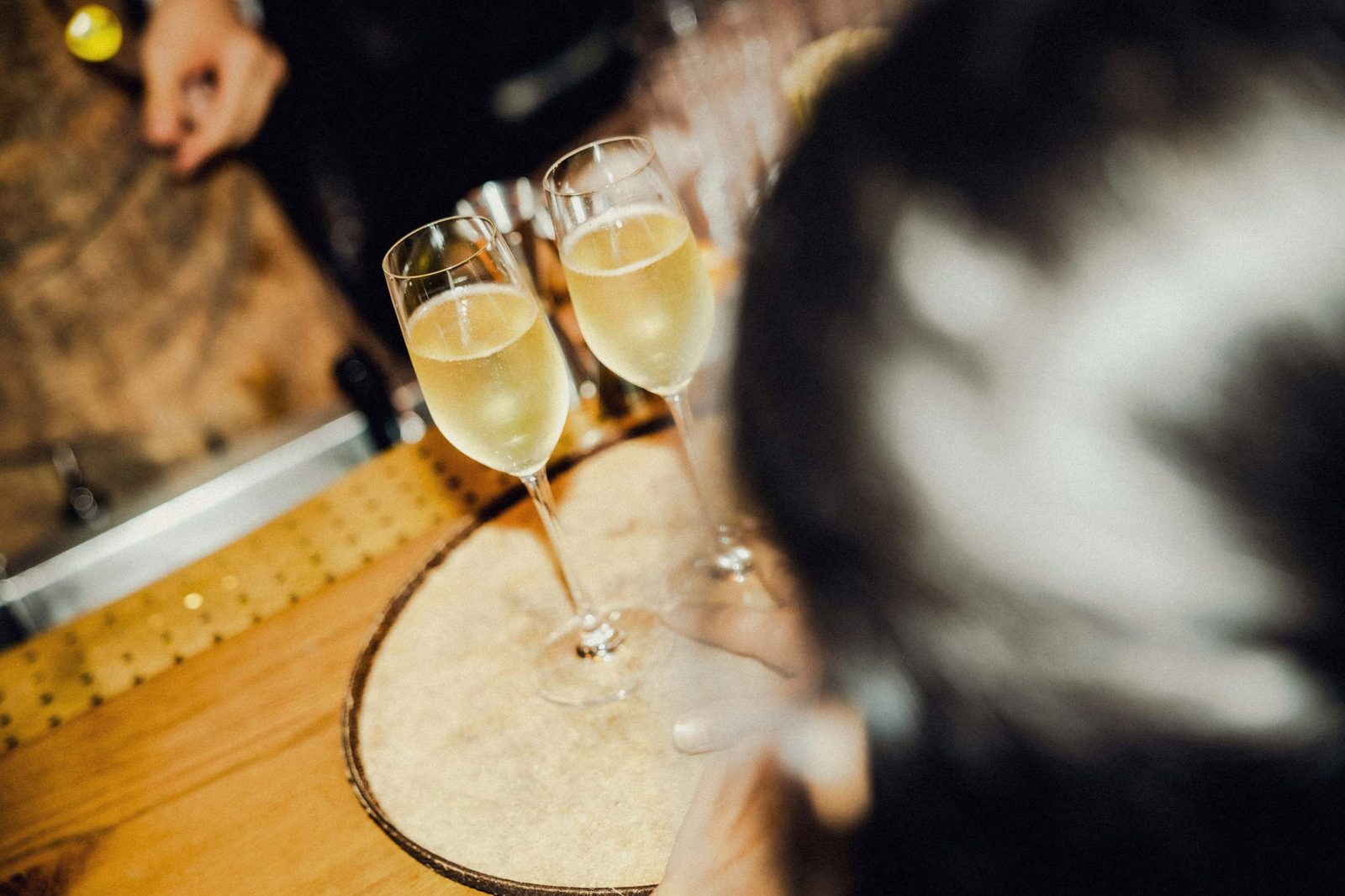 Two glasses of champagne sit on a round tray on a wooden bar, with a blurred person in the foreground and another persons hand visible in the background.