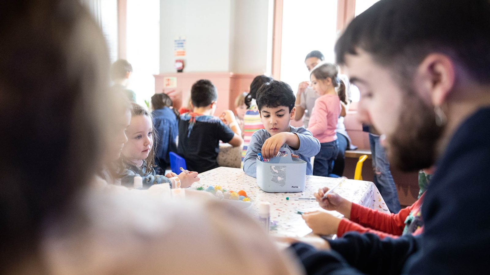 A group of children and adults sit around tables in a brightly lit classroom, engaged in arts and crafts activities with paper and colorful materials. One boy in the center focuses on his project.