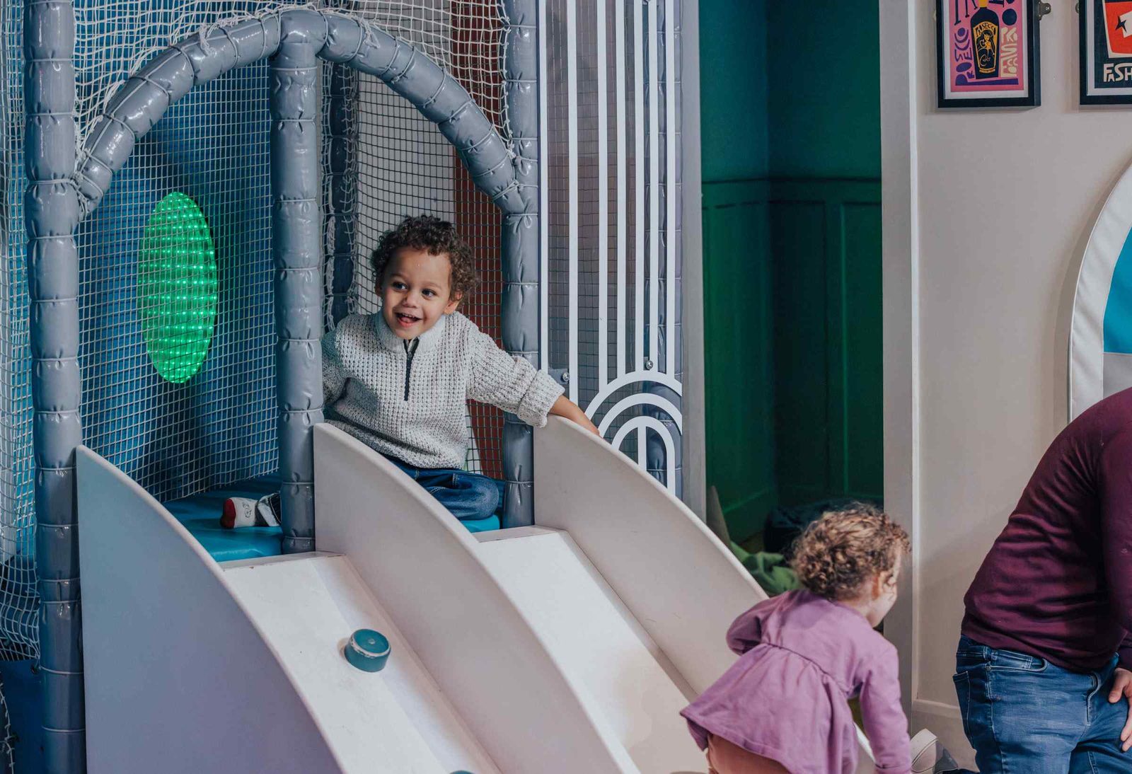 A young child smiles while sitting at the top of an indoor slide structure, and another child with curly hair plays nearby. The play area has netting, pastel colors, and playful decor on the walls.