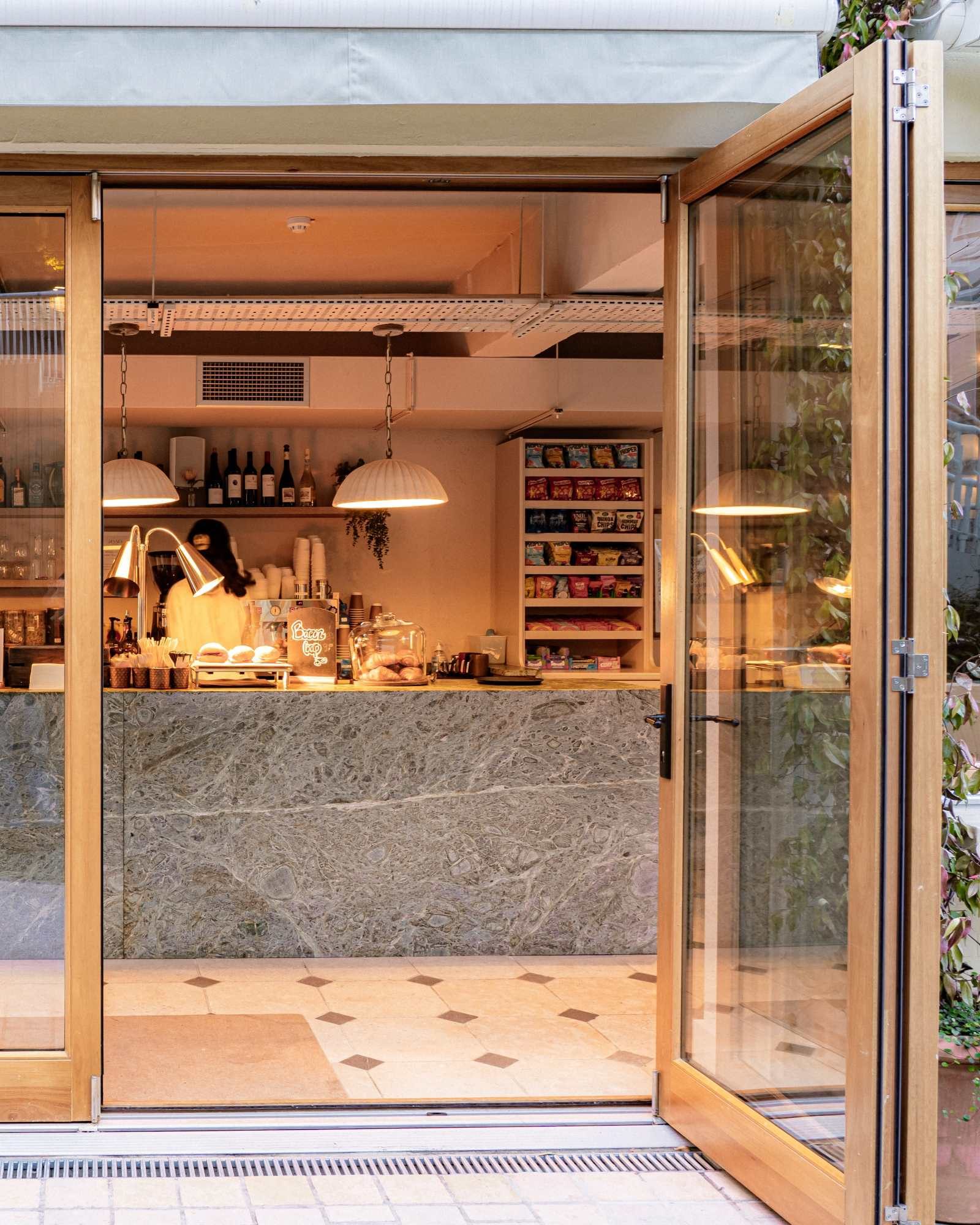 A cozy cafe with open glass doors, a marble counter, hanging lamps, and shelves stocked with snacks, drinks, and bottles. Warm lighting creates an inviting atmosphere inside.