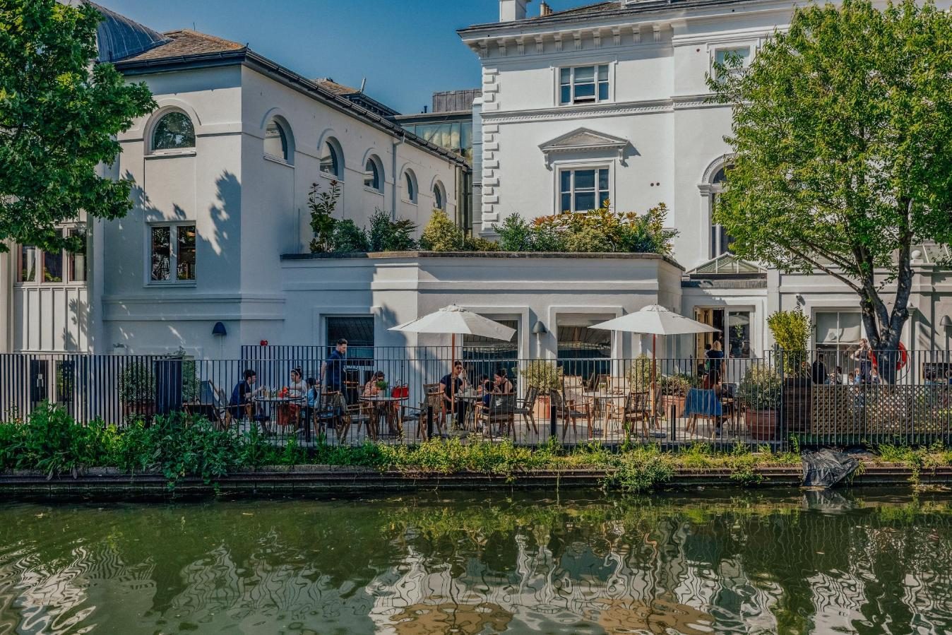 Outdoor café with people sitting under white umbrellas beside a canal, next to a white building with greenery and trees, on a sunny day.