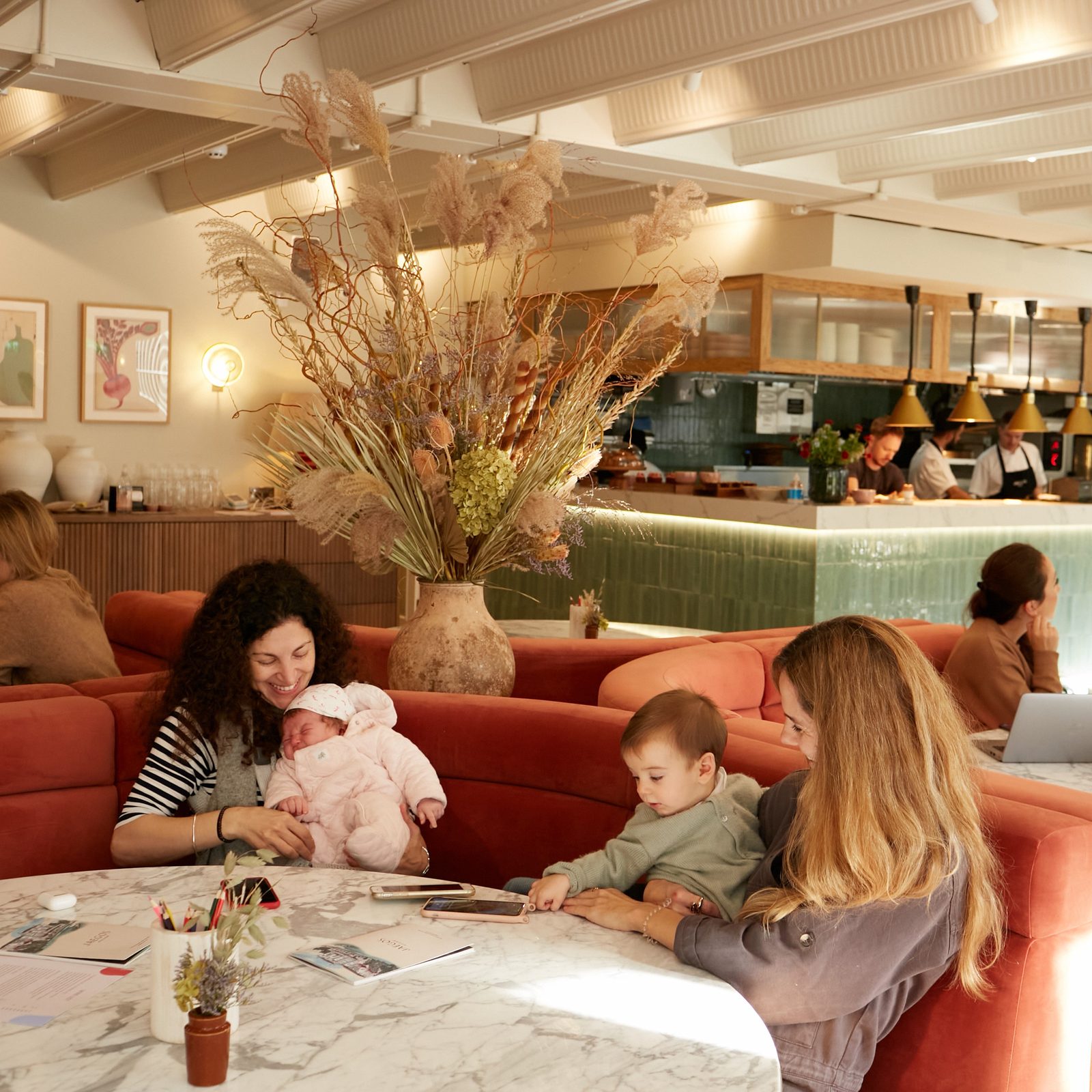 Two women sit at a marble table in a cozy cafe with a baby and a toddler. Dried flowers in a large vase decorate the table, and other people are seated in the background near the bar and green-tiled counter.