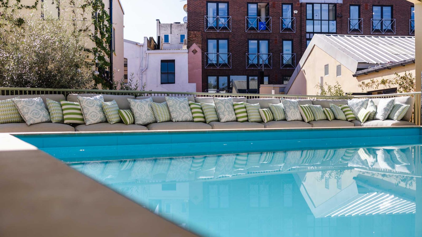 A modern outdoor pool with turquoise water, surrounded by a long cushioned bench lined with green and white patterned pillows. Urban buildings are visible in the background under a clear sky.