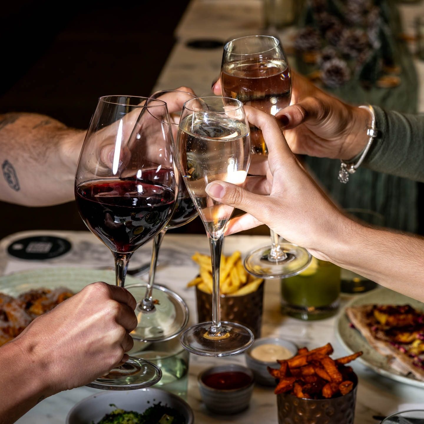 Four hands clinking wine and champagne glasses over a table with plates of food, fries, and dipping sauces, creating a celebratory and festive atmosphere.