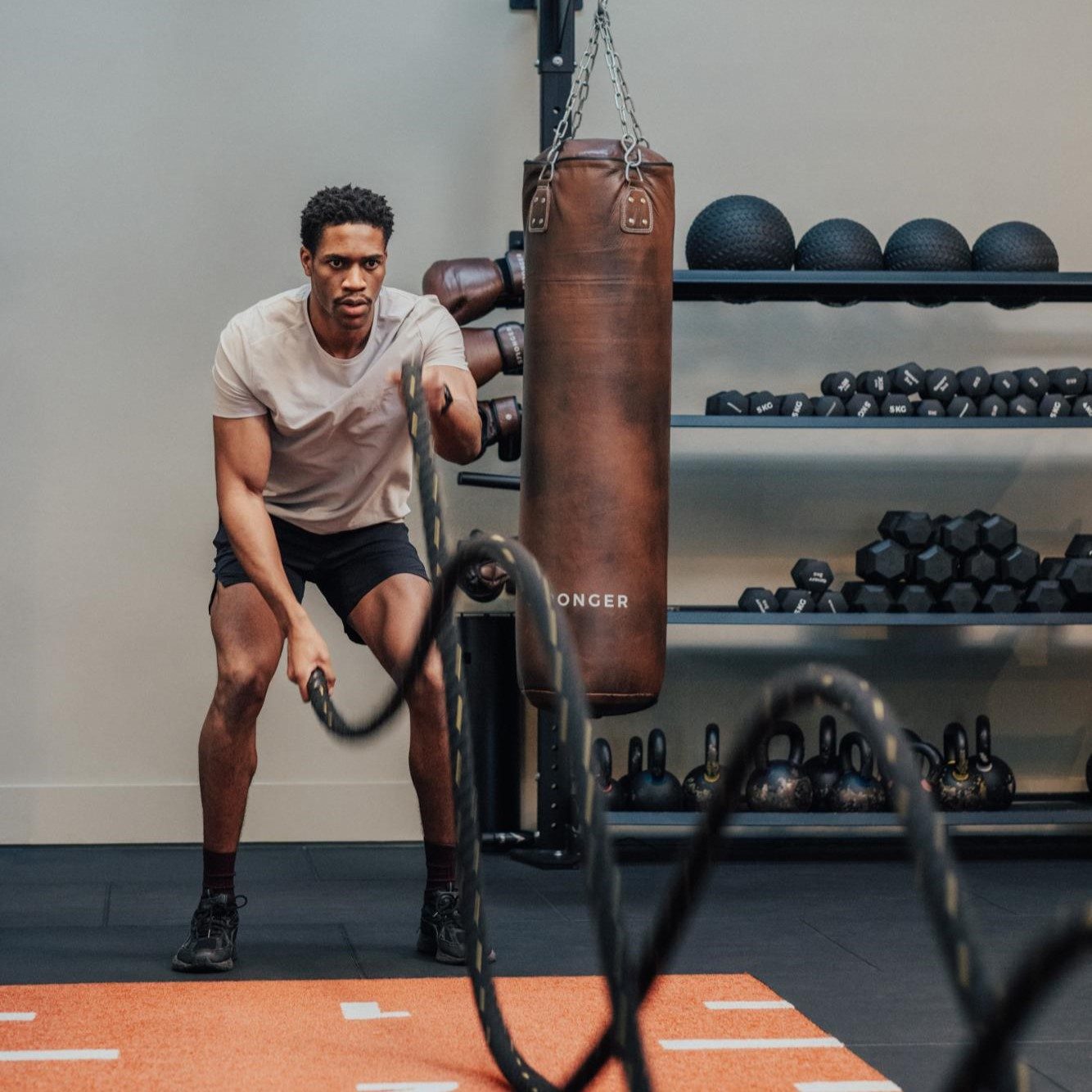 A man in athletic wear exercises with battle ropes in a gym, with a punching bag, kettlebells, dumbbells, and medicine balls visible in the background.