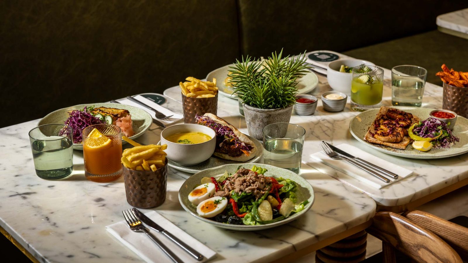 A marble table set with plates of salad, fries, soup, sandwiches, and pizza, accompanied by drinks and potted plants, arranged for a meal in a cozy restaurant setting.
