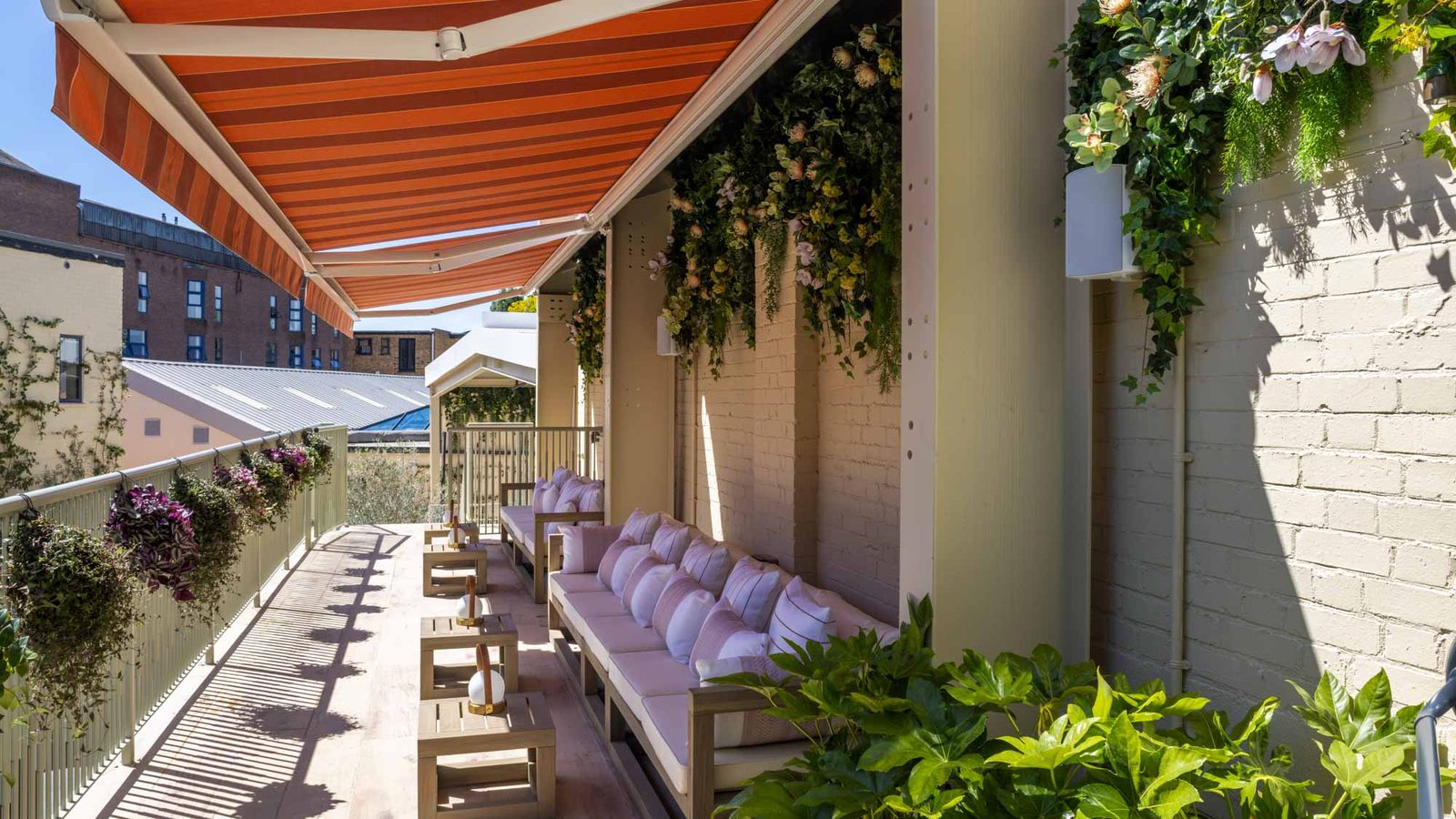 A sunlit outdoor patio with cushioned seating, wooden tables, hanging plants, and a red-and-white striped awning, overlooking nearby buildings and other greenery.