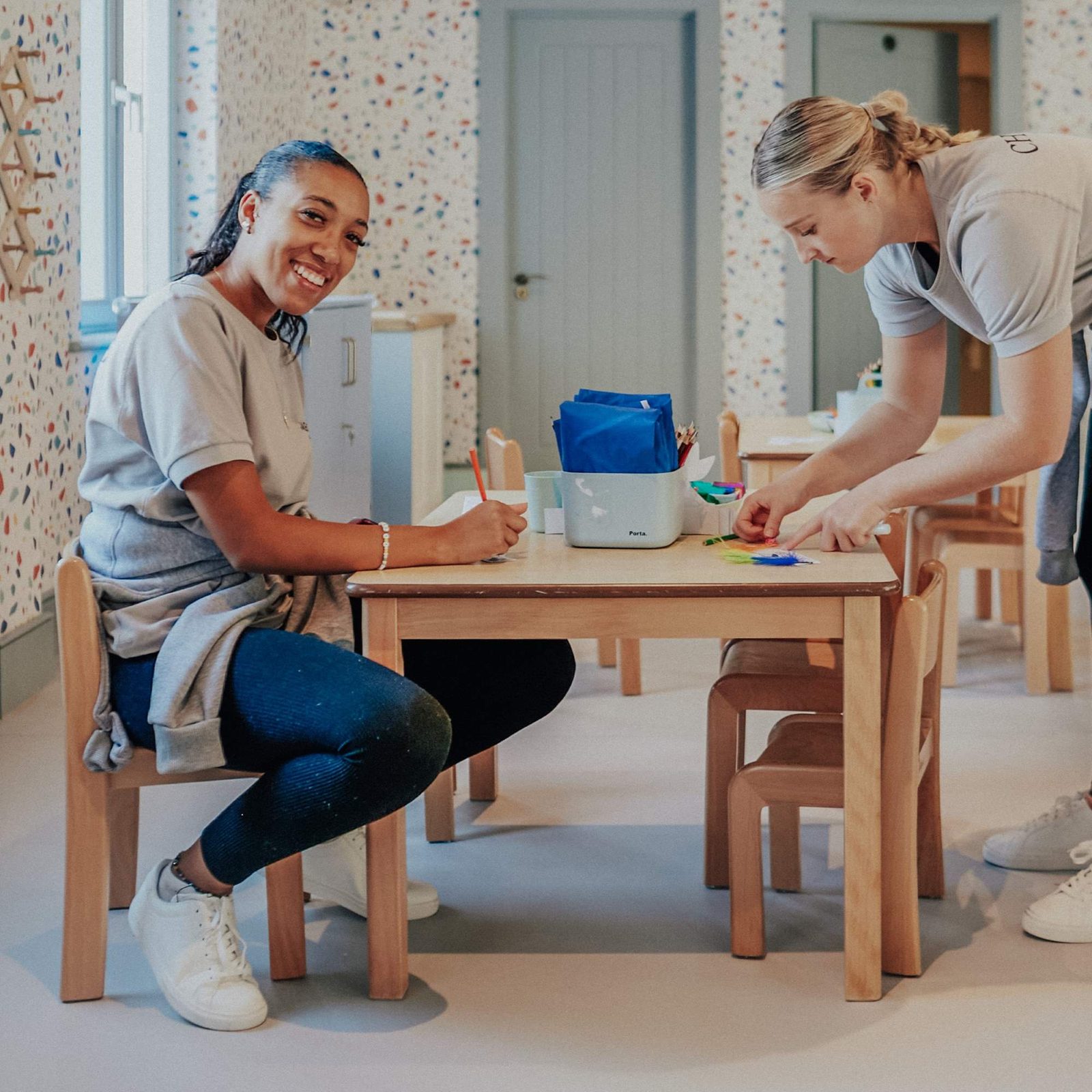 Two women work together at a small wooden table in a colorful, child-friendly classroom. One woman sits and smiles at the camera while writing; the other stands and arranges materials on the table.