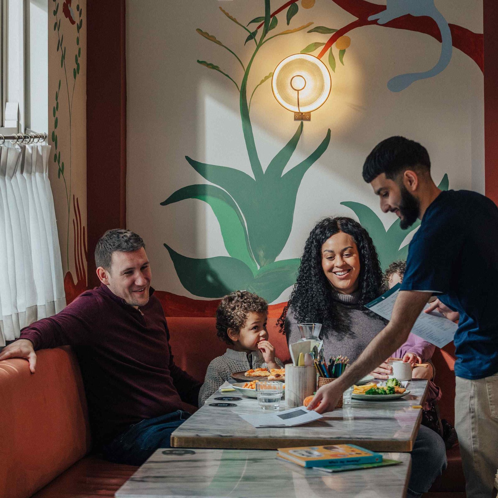 A family of four sits at a restaurant booth, smiling as a waiter hands them the bill. Two children sit between the parents, with meals on the table. The wall behind them has decorative plant artwork and a round light fixture.