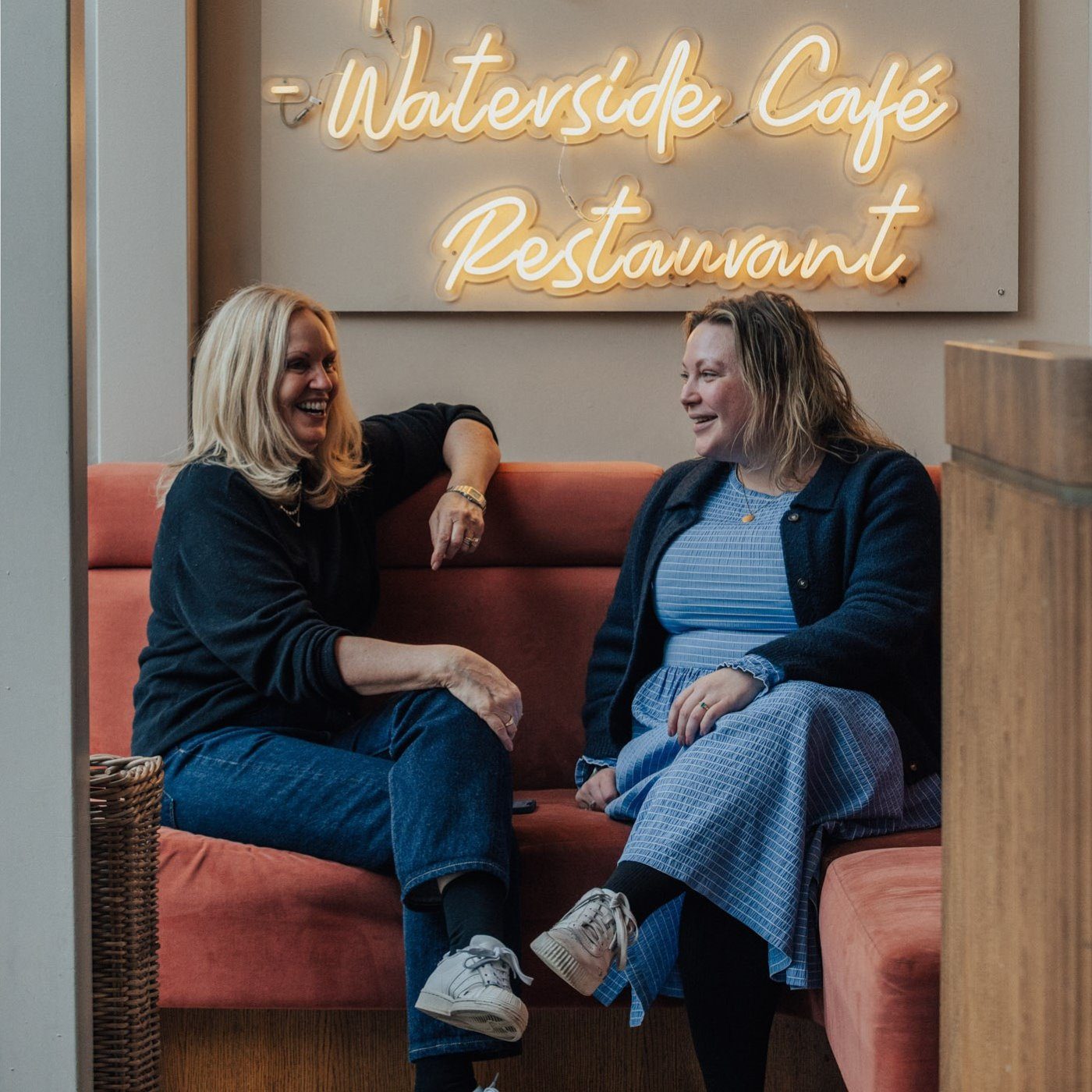 Two women sit on a peach-colored bench, smiling and chatting, beneath a neon sign that reads Waterside Café Restaurant. The setting appears cozy and welcoming.
