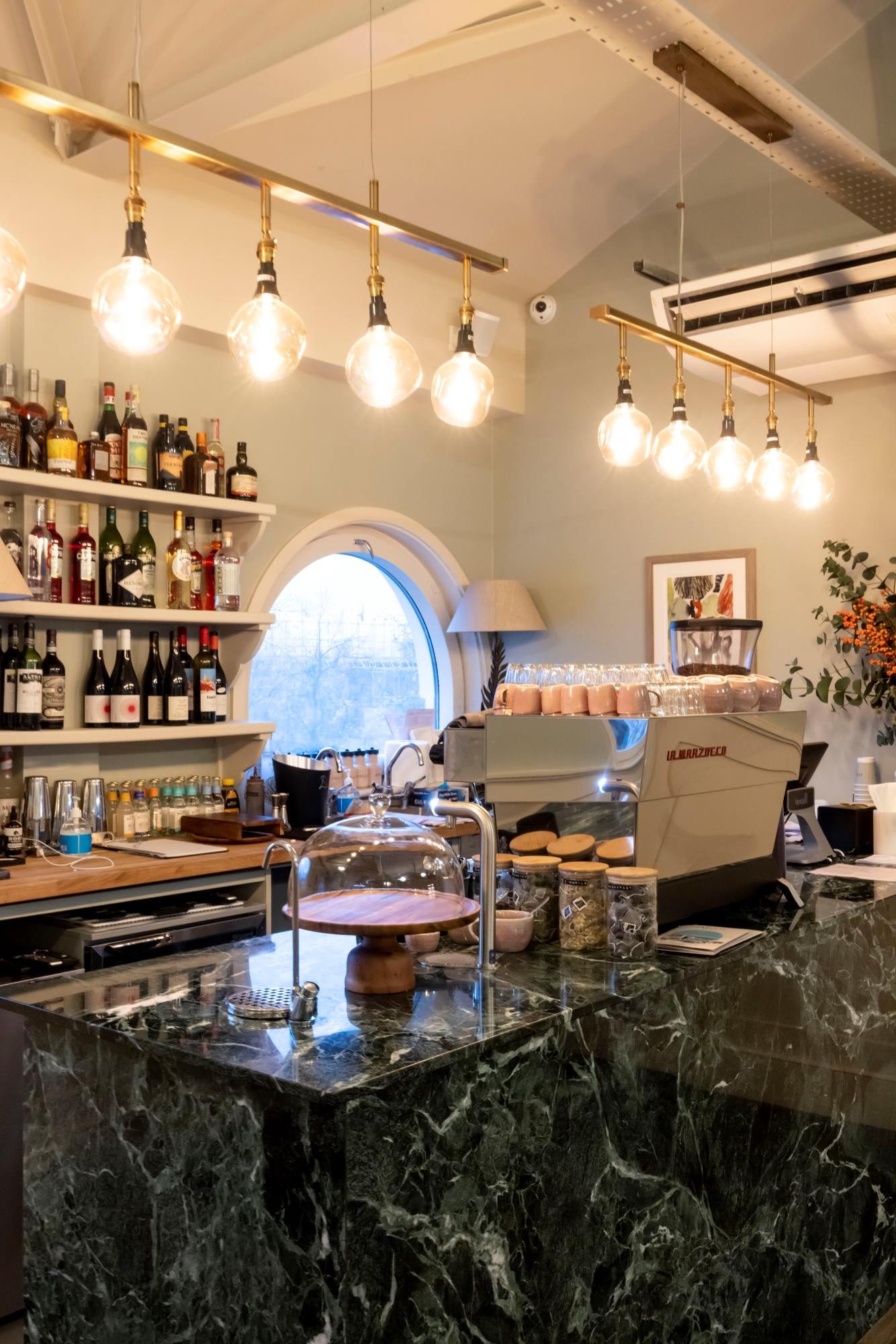 A cozy café counter with a green marble surface, espresso machine, pastries under glass domes, shelves of liquor bottles, hanging pendant lights, and a round window in the background.