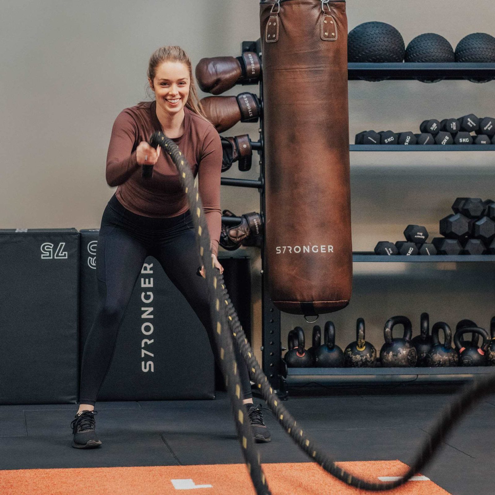 A woman in athletic wear exercises with battle ropes in a gym, smiling. There are kettlebells, dumbbells, medicine balls, and a punching bag visible in the background.