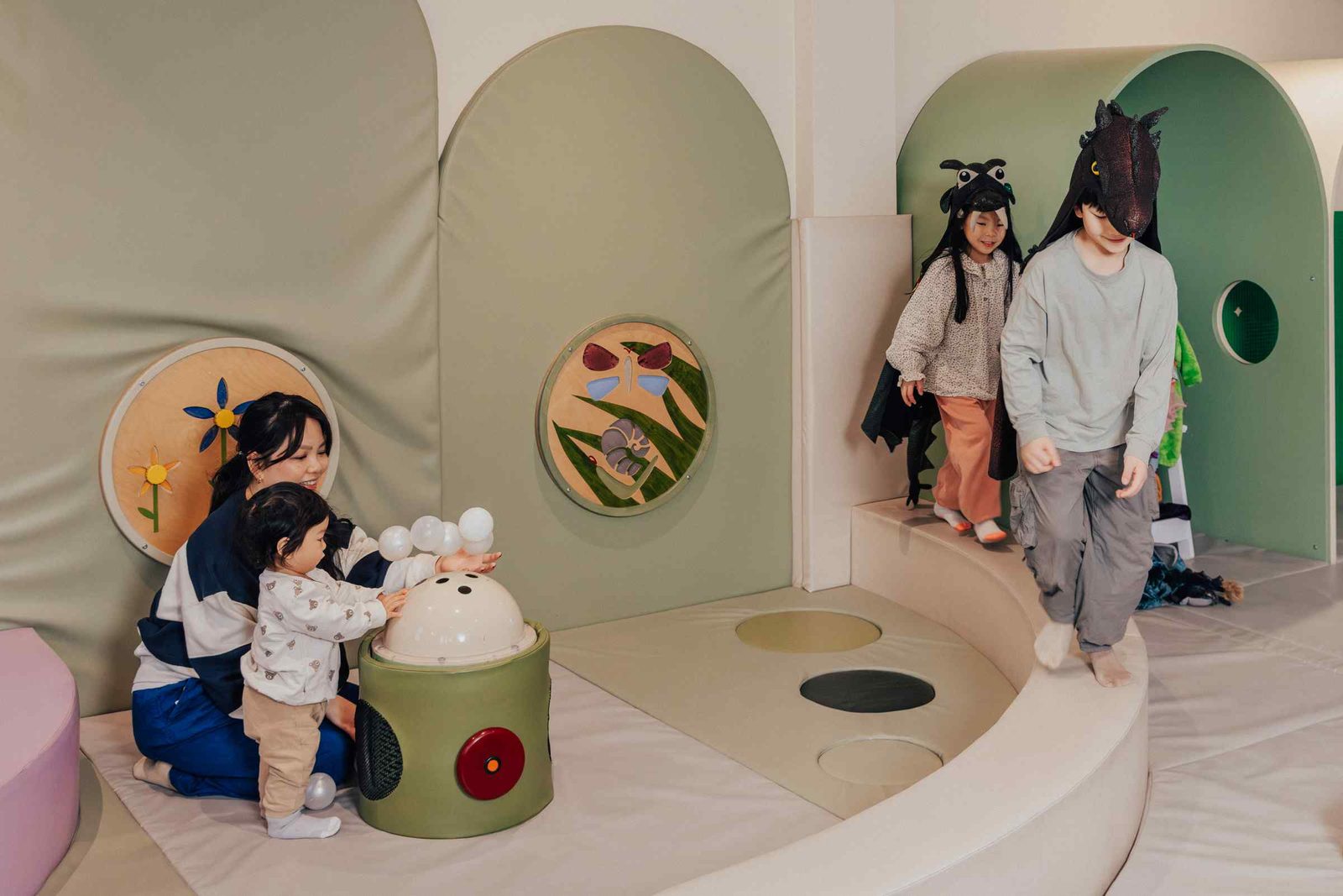 Children play in a soft, indoor play area; one child presses a bubble-making toy with an adult, while two kids in animal costumes walk nearby, all surrounded by pastel-colored padded walls and playful decorations.