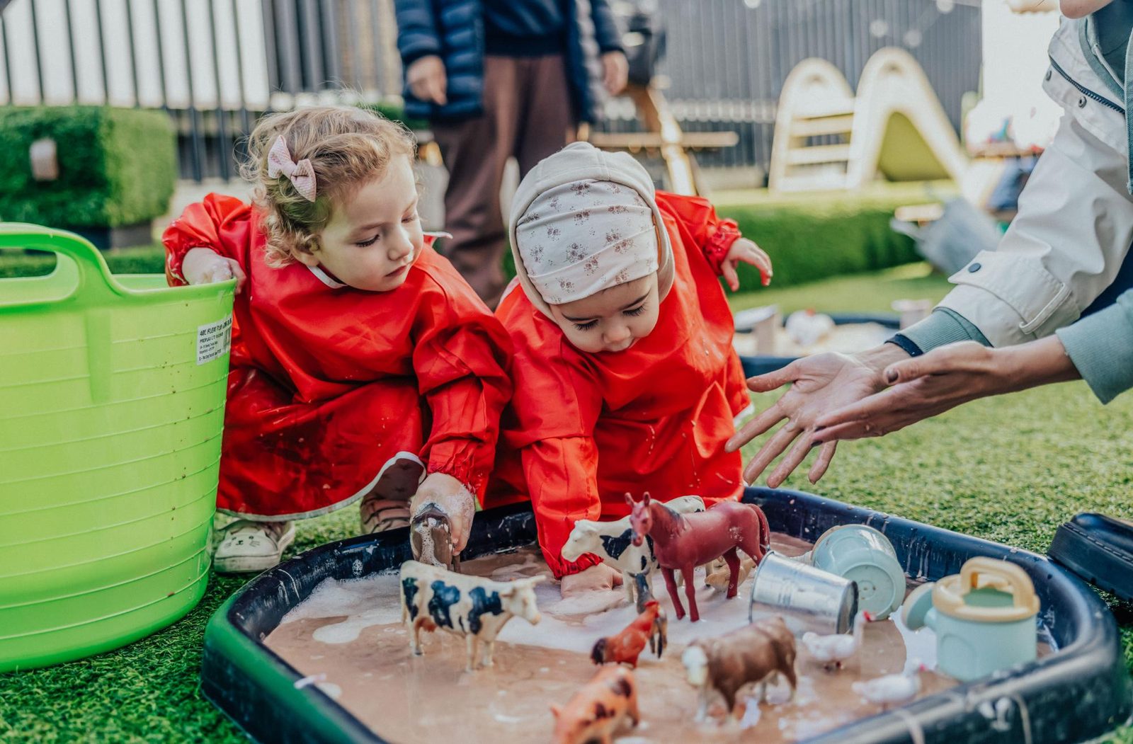 Two young children in red smocks play with toy farm animals and sensory materials in a black tray outdoors, while an adult’s hand reaches in to help. The scene is playful and takes place on artificial grass.