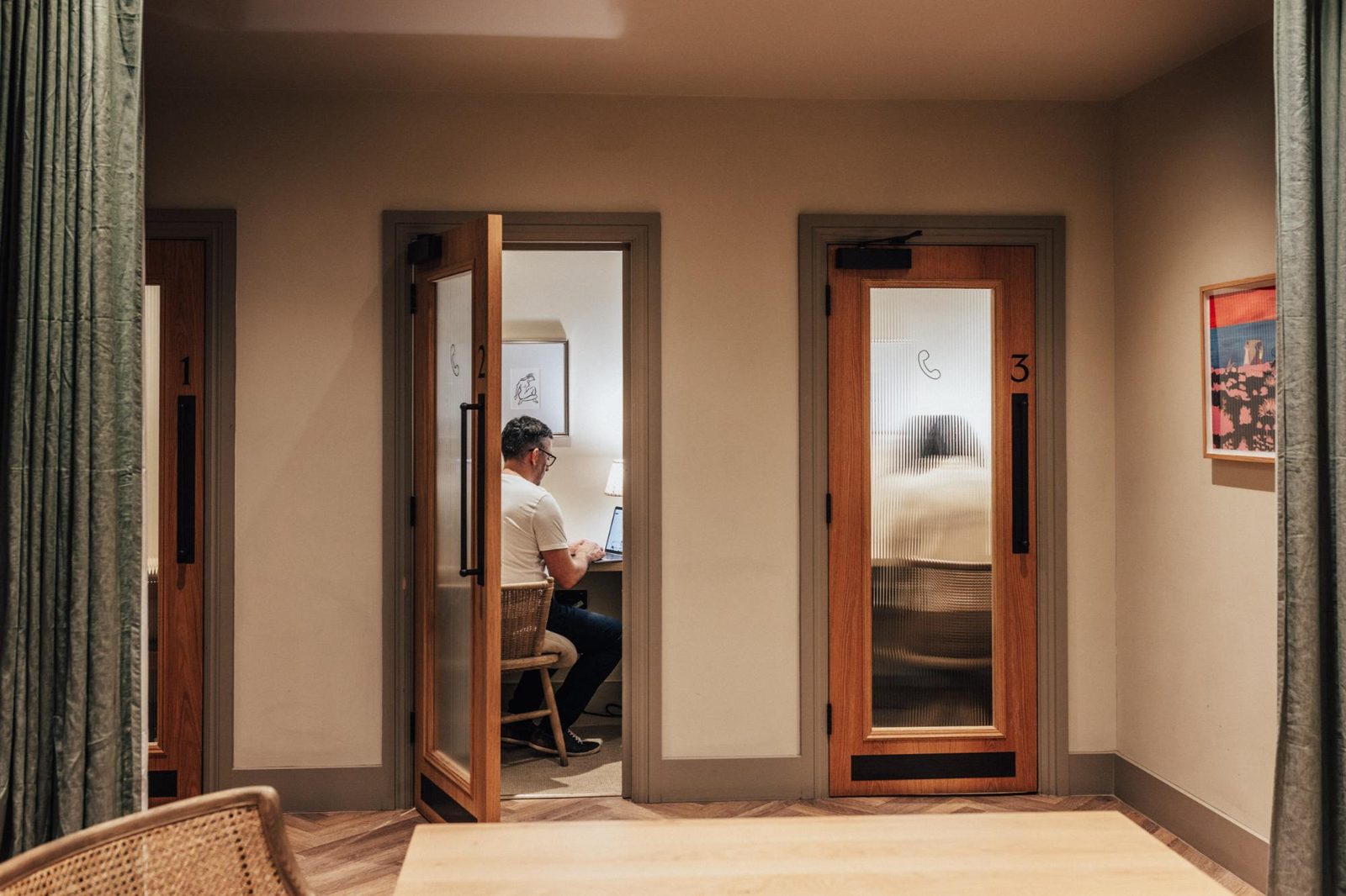 A man sits at a desk working on a laptop inside a small office booth with an open door, while another adjacent booth has a person inside who appears blurred in motion behind a glass door.