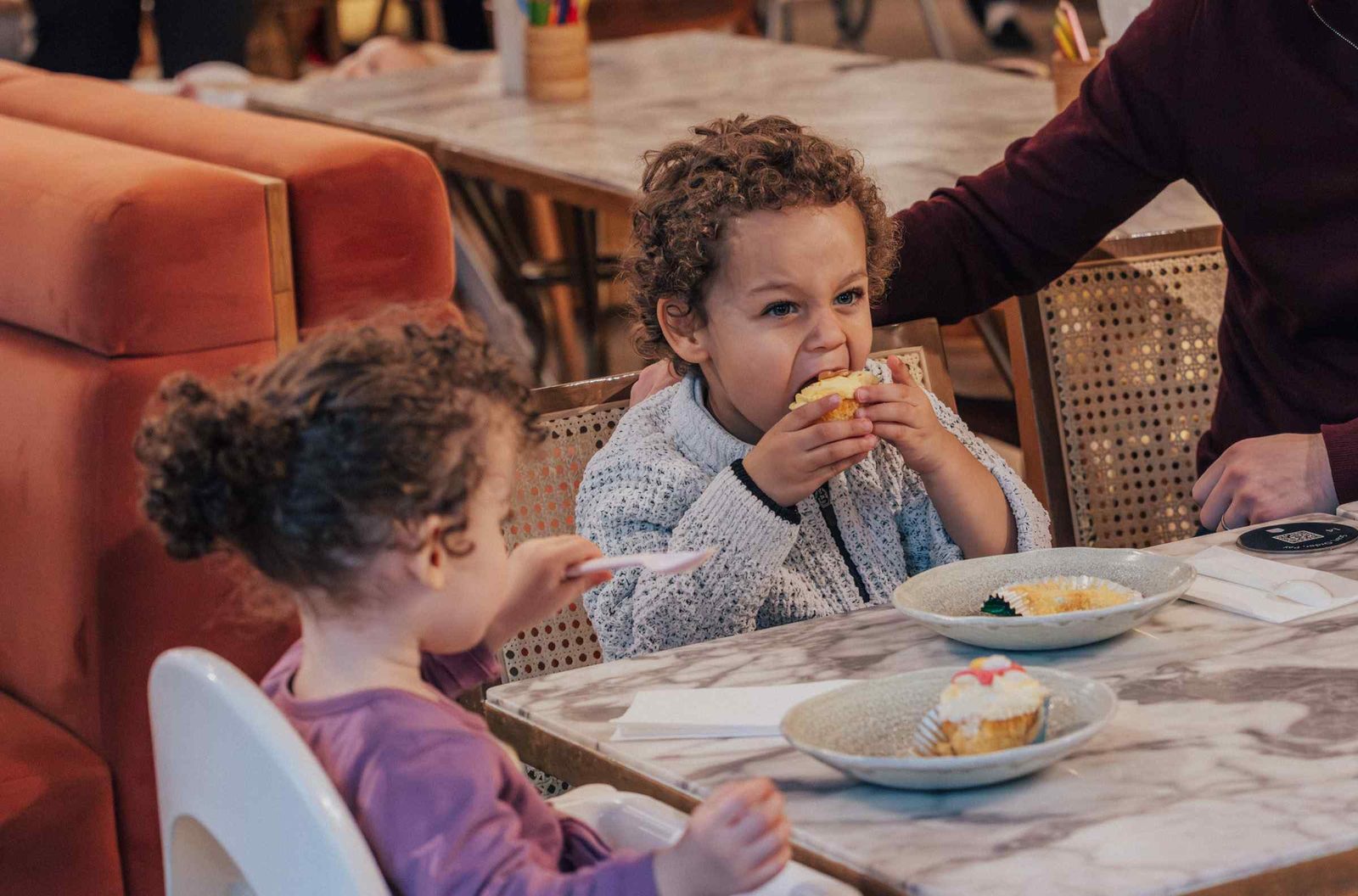 Two young children sit at a marble table in a restaurant. One child enthusiastically bites into a pastry, while the other looks at a cupcake. An adults arm rests on a chair nearby. Plates and napkins are on the table.