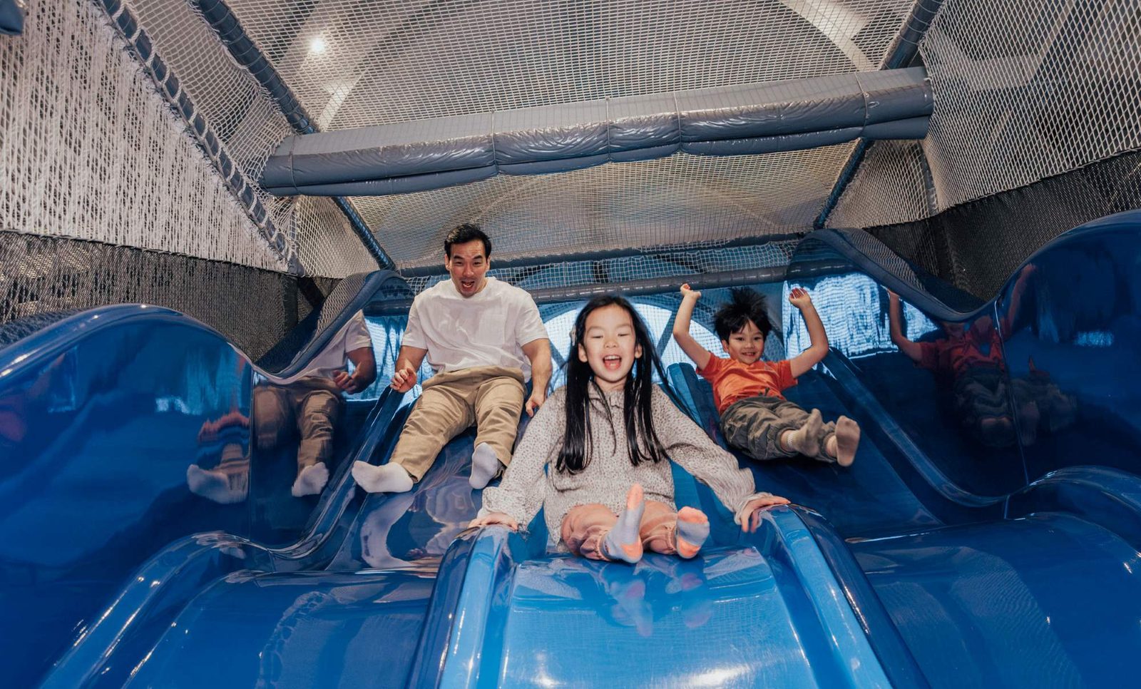 A man and two children are sliding down bright blue slides indoors, smiling and having fun. The background shows netting and play structures, suggesting they are in an indoor playground.