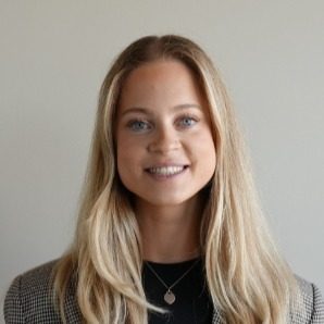 A young woman with long blonde hair, wearing a checkered blazer and a black top, smiles at the camera against a plain light-colored background.