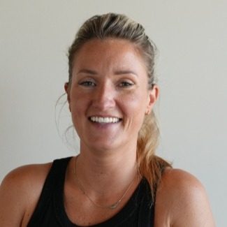 A woman with light brown hair pulled back, wearing a black sleeveless top and a delicate necklace, smiling in front of a plain light-colored background.