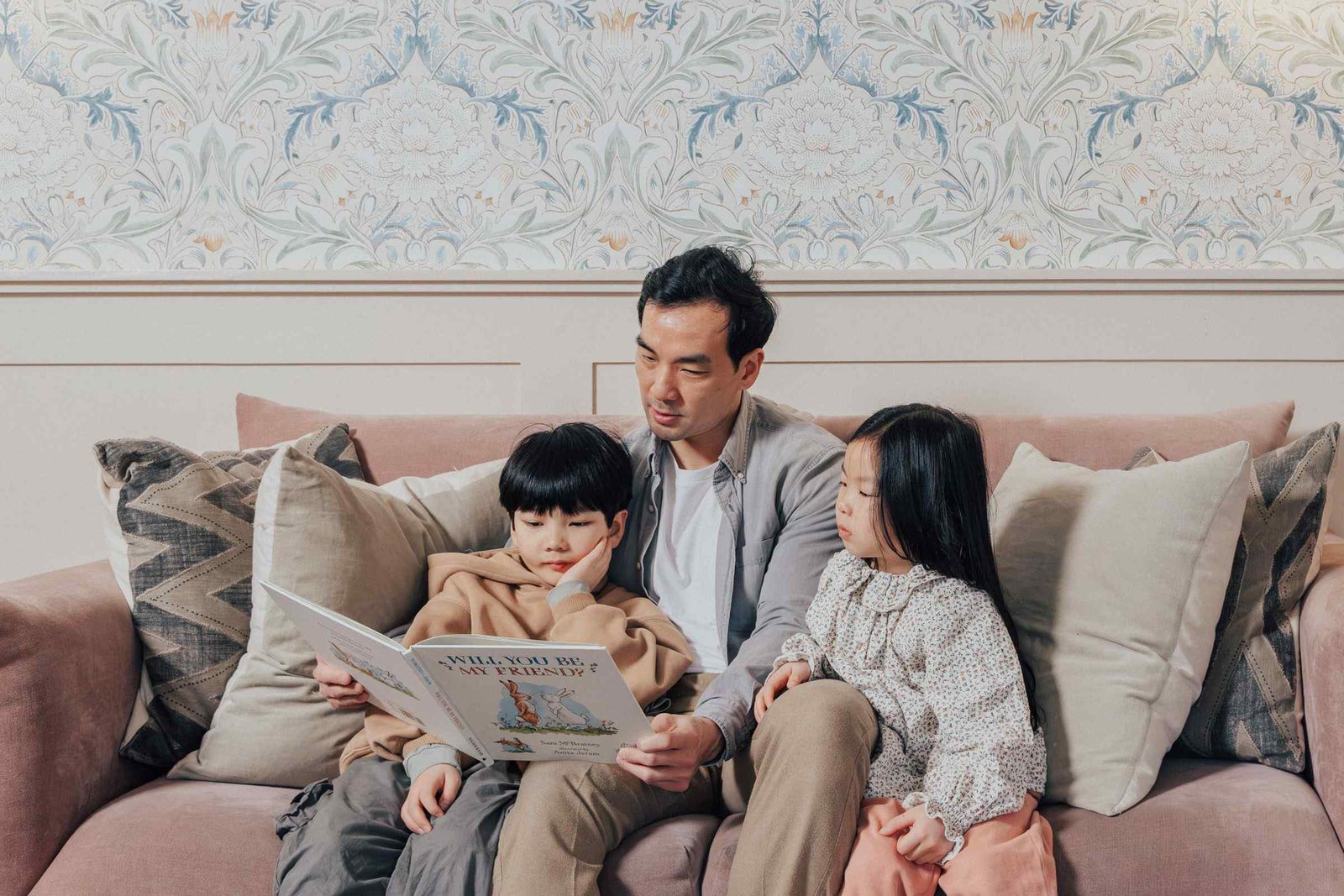 A man sits on a sofa reading a picture book to a young boy and girl. All three are surrounded by cushions, and the background features a decorative floral wallpaper.