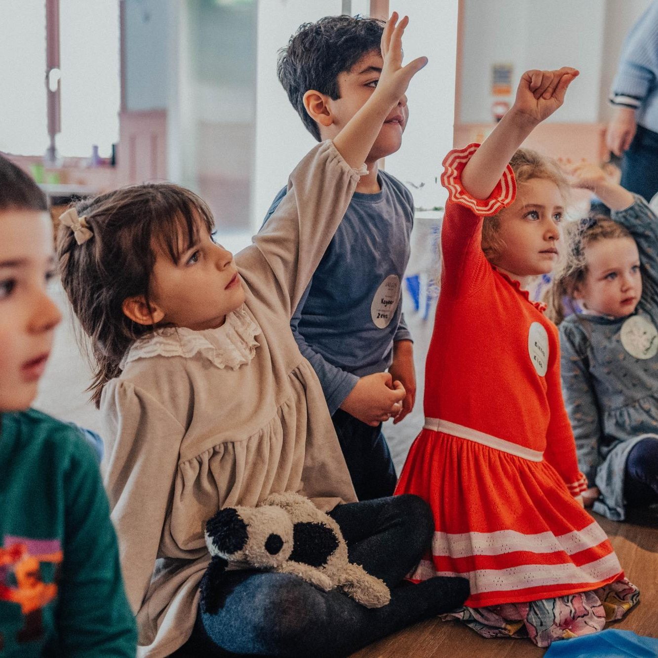 A group of young children sitting on the floor in a classroom. Two children in front, one girl in a red dress and one boy, have their hands raised. A stuffed panda is on the girls lap. Other kids watch attentively.