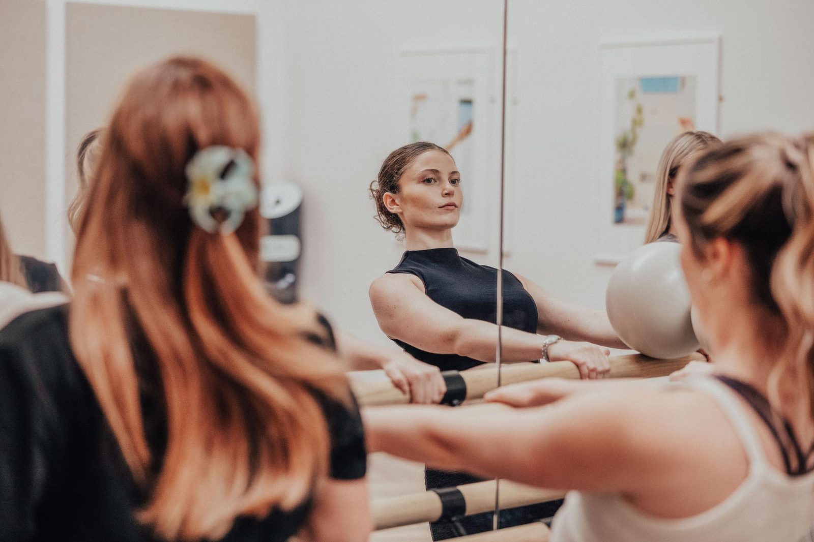 Three women exercise at a ballet barre in a studio, focusing on their form in a mirrored wall. One woman, centered and facing the mirror, holds a neutral expression while balancing a gray exercise ball.