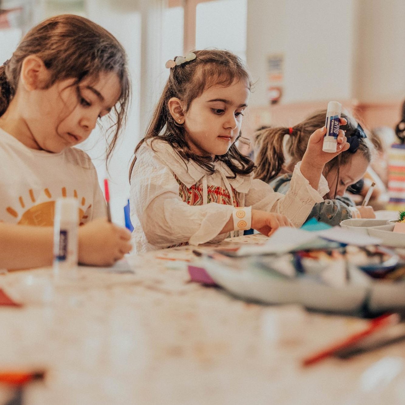 Three young children sit at a table doing arts and crafts. One girl is drawing, while another holds up a glue stick, both focused on their creative projects with various materials spread out in front of them.