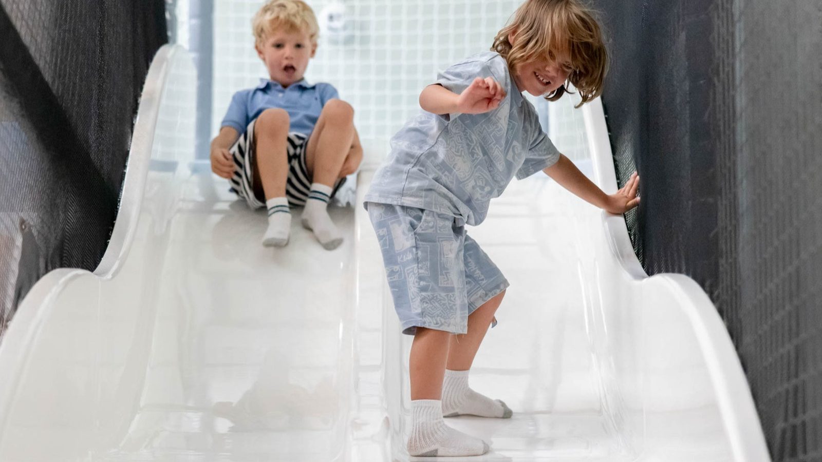 Two young children play on a shiny white slide. One child stands and holds the side while sliding down, while the other sits at the top, preparing to slide. Both are barefoot and wearing casual clothes.
