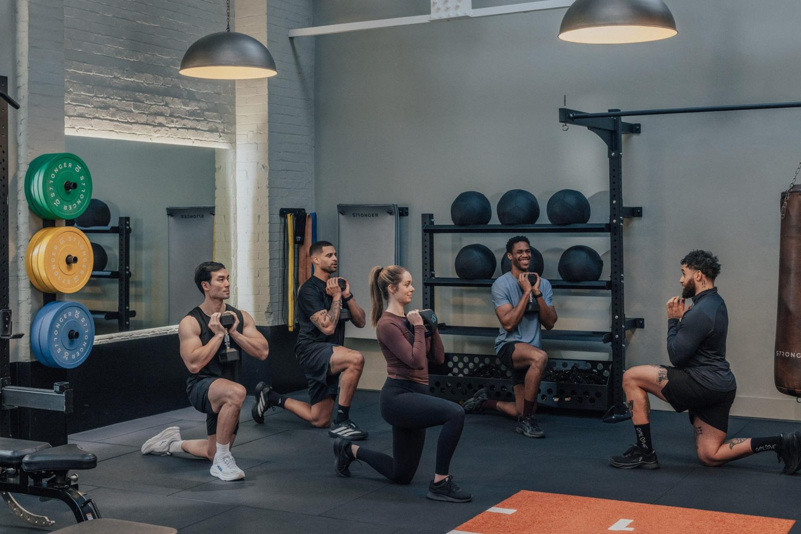 Five people in athletic wear perform weighted lunges together in a gym, following an instructor. Weight plates, medicine balls, and gym equipment are visible in the background.