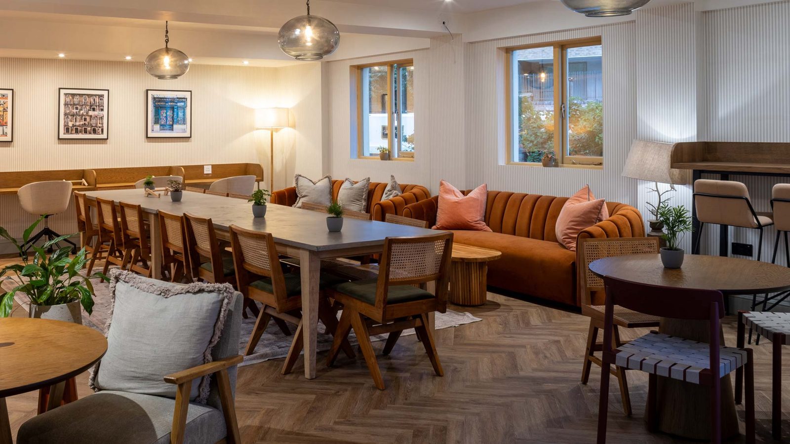 A cozy, modern lounge with wooden tables and chairs, an orange sofa with pillows, soft lighting from ceiling lamps, and large windows. Plants and framed pictures add warmth to the space, and a bar area is visible on the right.