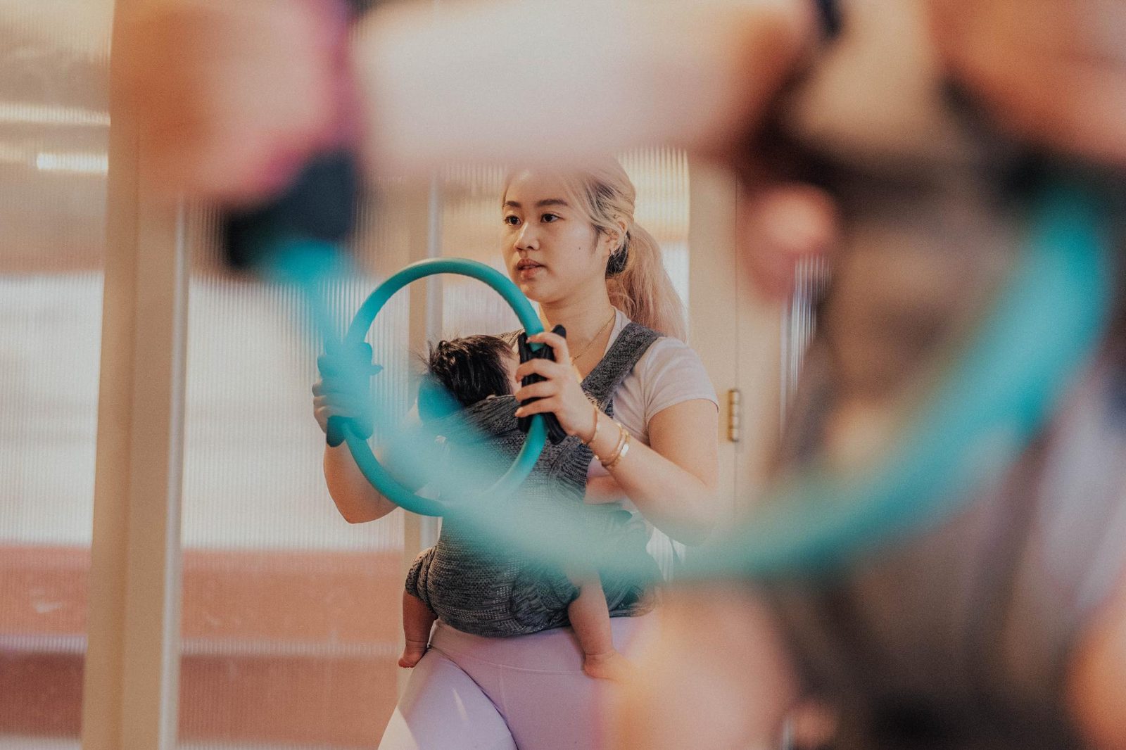 A woman wearing workout clothes holds a fitness ring while carrying a baby in a front carrier. She appears focused on her exercise, and her reflection can be seen in a mirror.